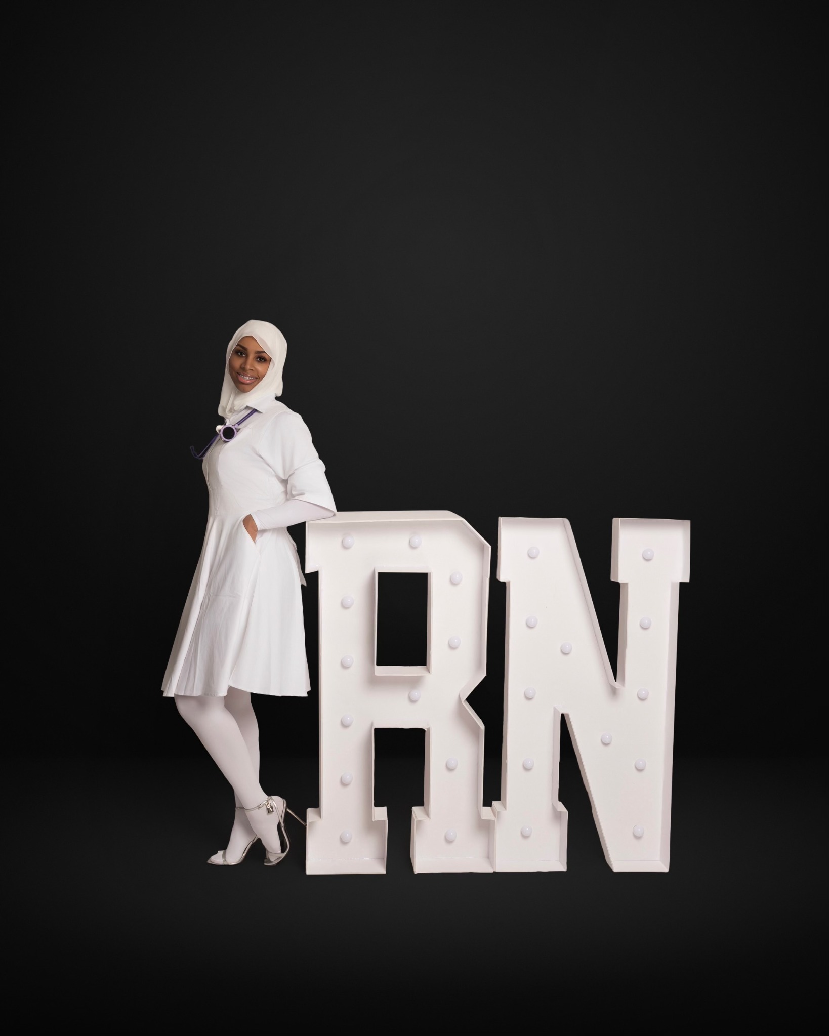 Ny’Rossha Alim in white nursing attire standing beside large RN letters